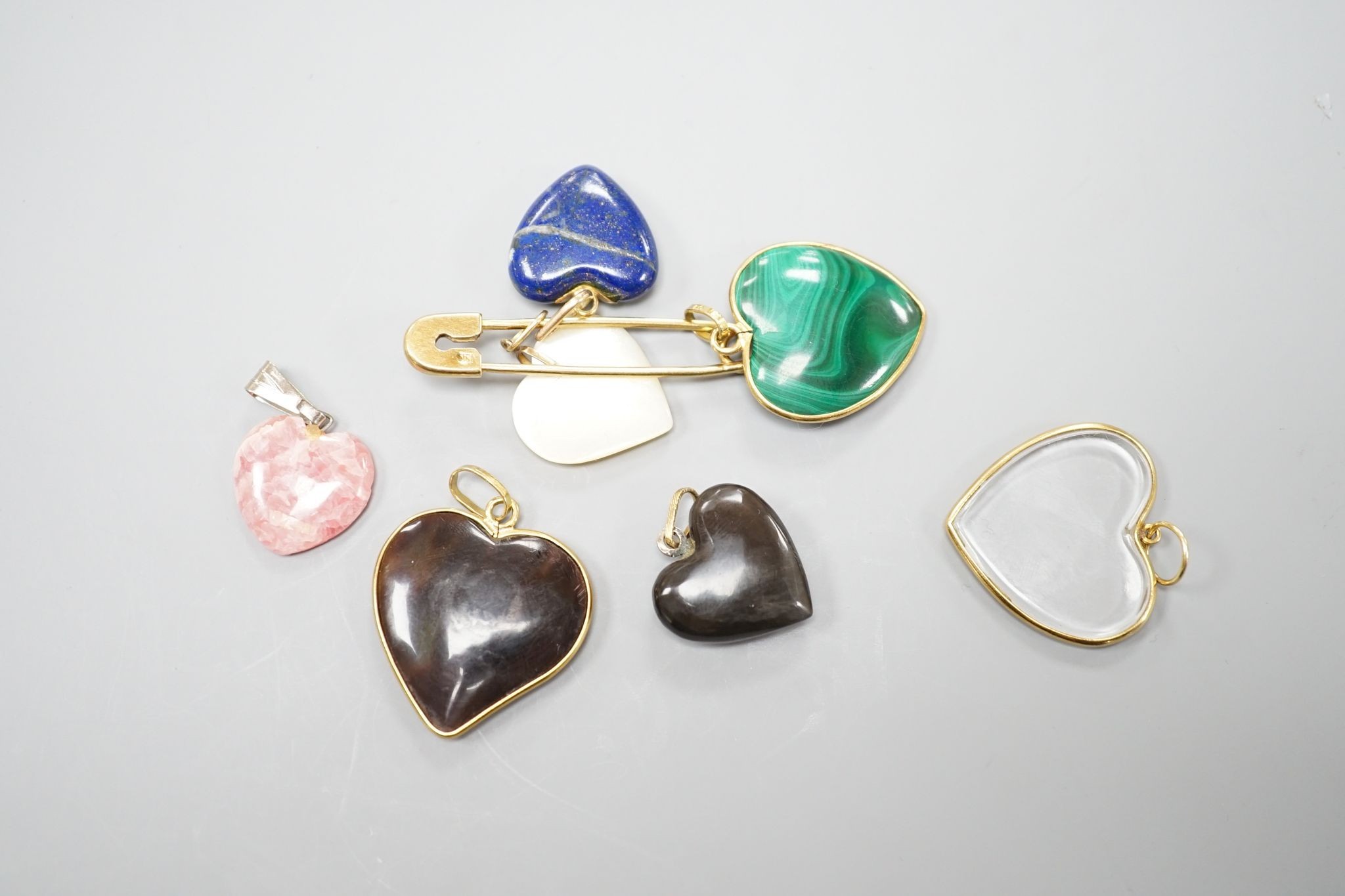 An 18ct safety pin, 47mm, hung with seven assorted heart shaped hardstone pendants or charms, two with Italian 750 yellow metal mounts.